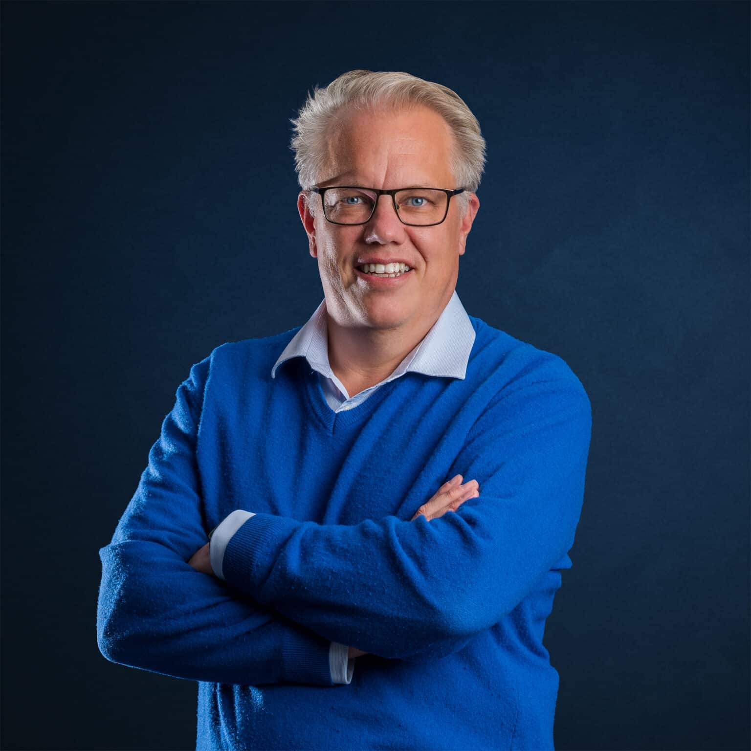 Mann mit Brille und blauer Pullover steht mit verschränkten Armen vor einem dunklen Hintergrund.