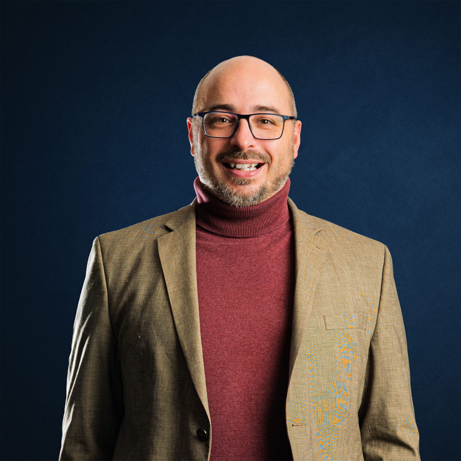 Mann mit Brille und Glatze trägt einen braunen Blazer über einem roten Rollkragenpullover vor einem blauen Hintergrund.