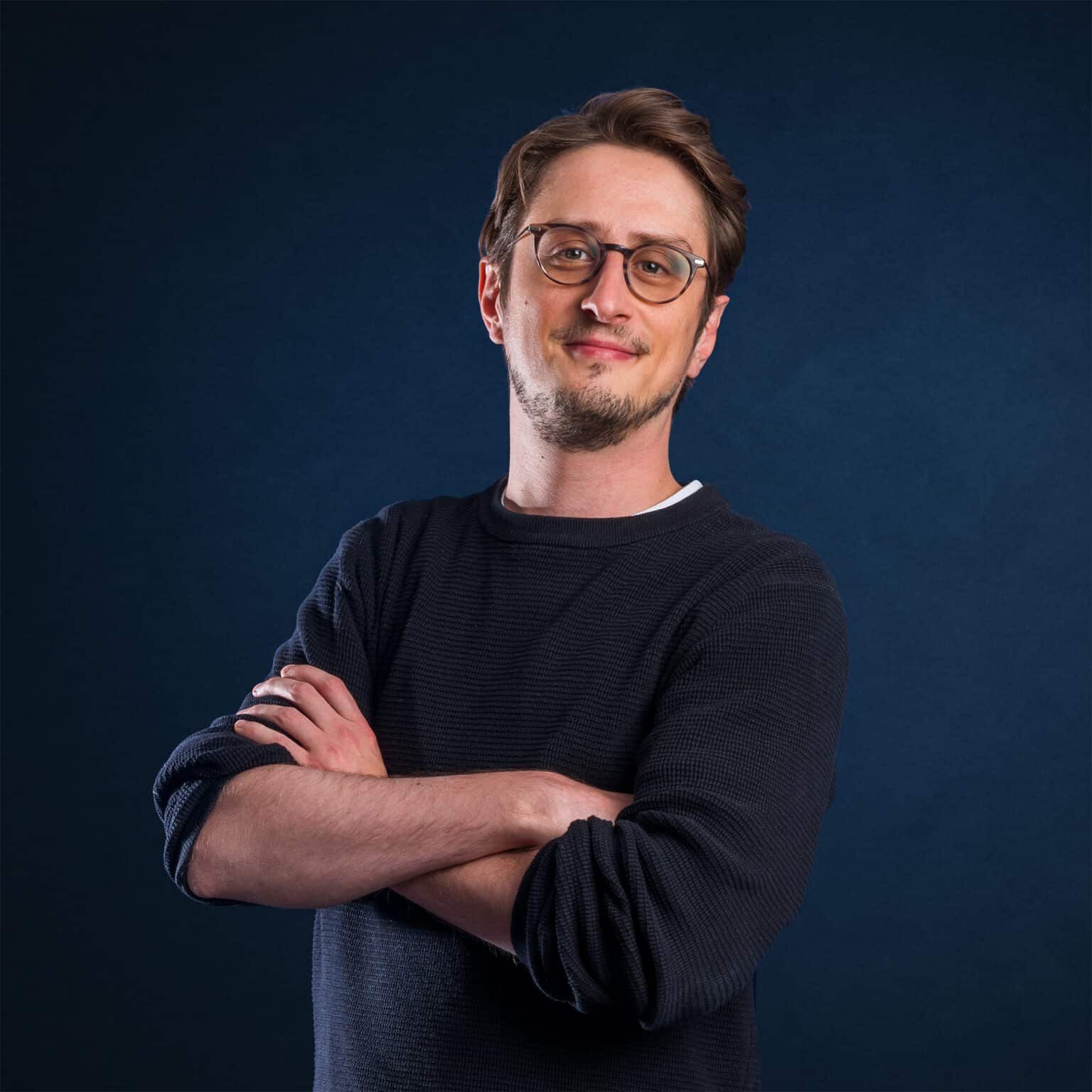 Philipp Hunold mit Brille und verschränkten Armen vor einem blauen Hintergrund.