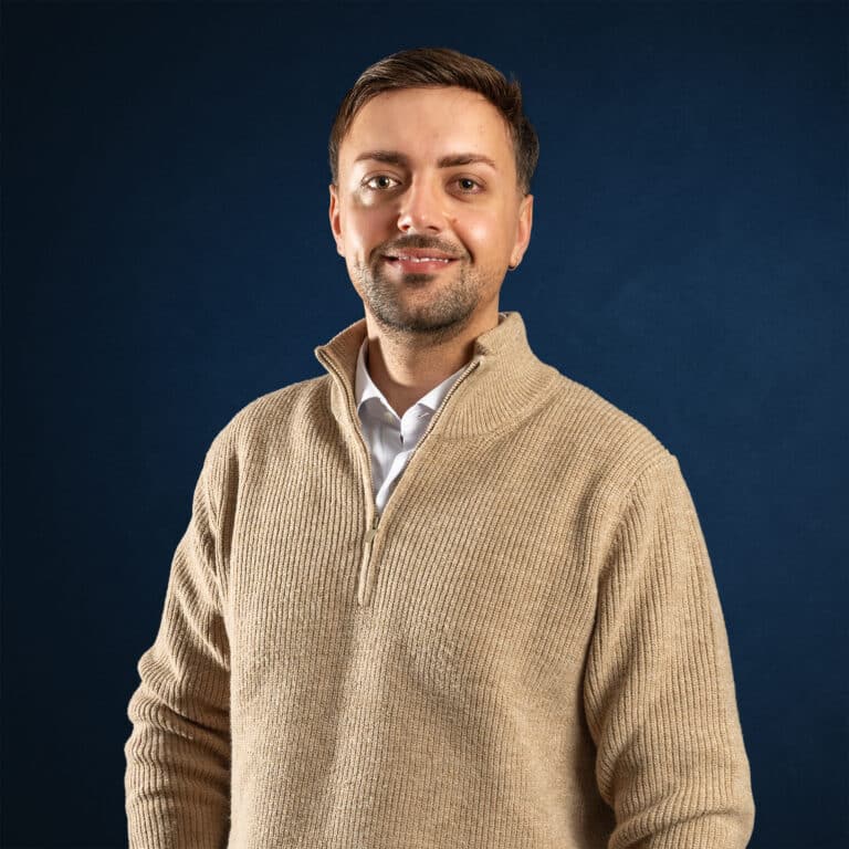 Dejan Rakanovic lächelt in einem beigen Strickpullover über einem weißen Hemd vor dunkelblauem Hintergrund, mit kurzem Bart und Ohrstecker.