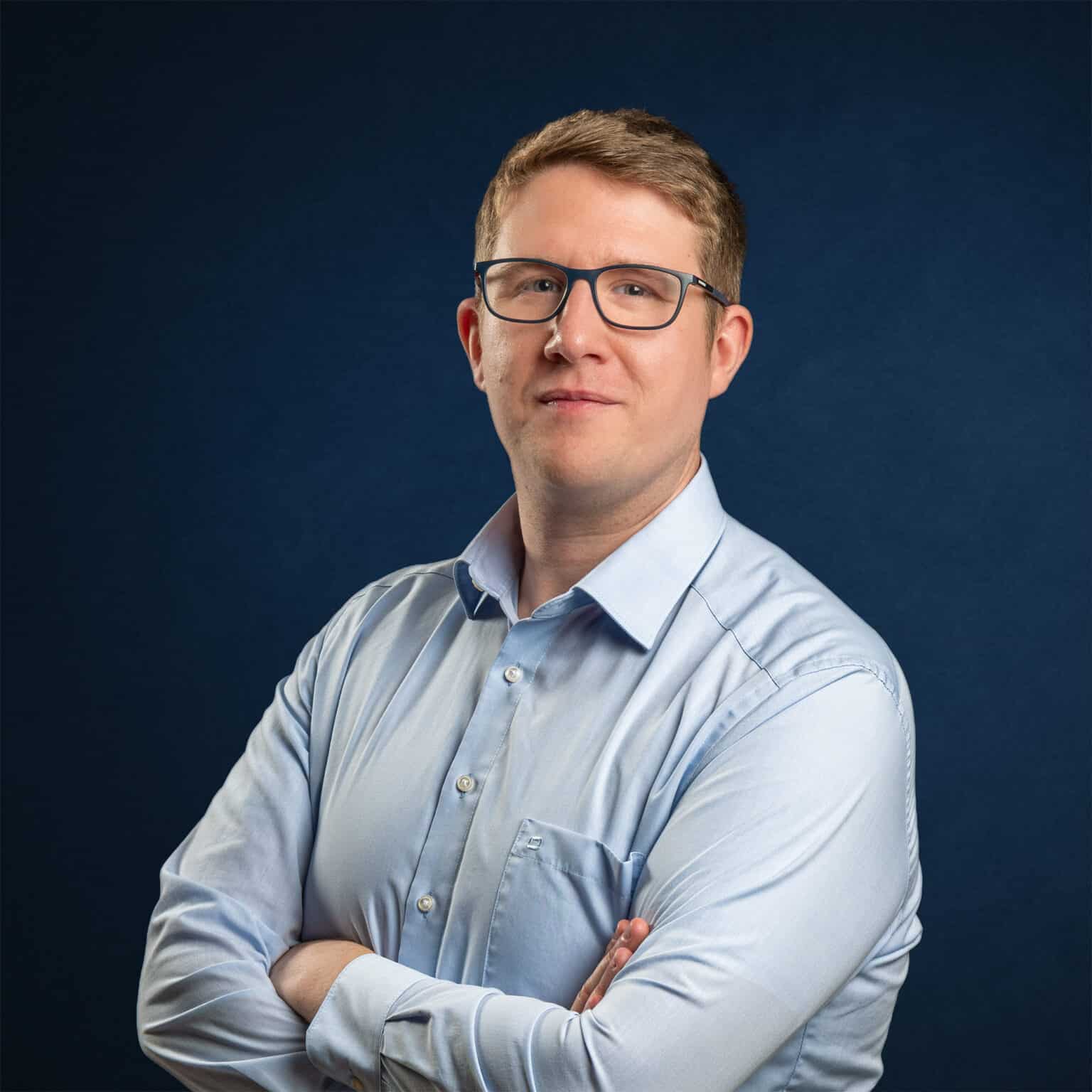 Andreas Klotz, LL.M. blickt mit Brille und verschränkten Armen in einem hellblauen Hemd vor dunkelblauem Hintergrund ruhig in die Kamera.