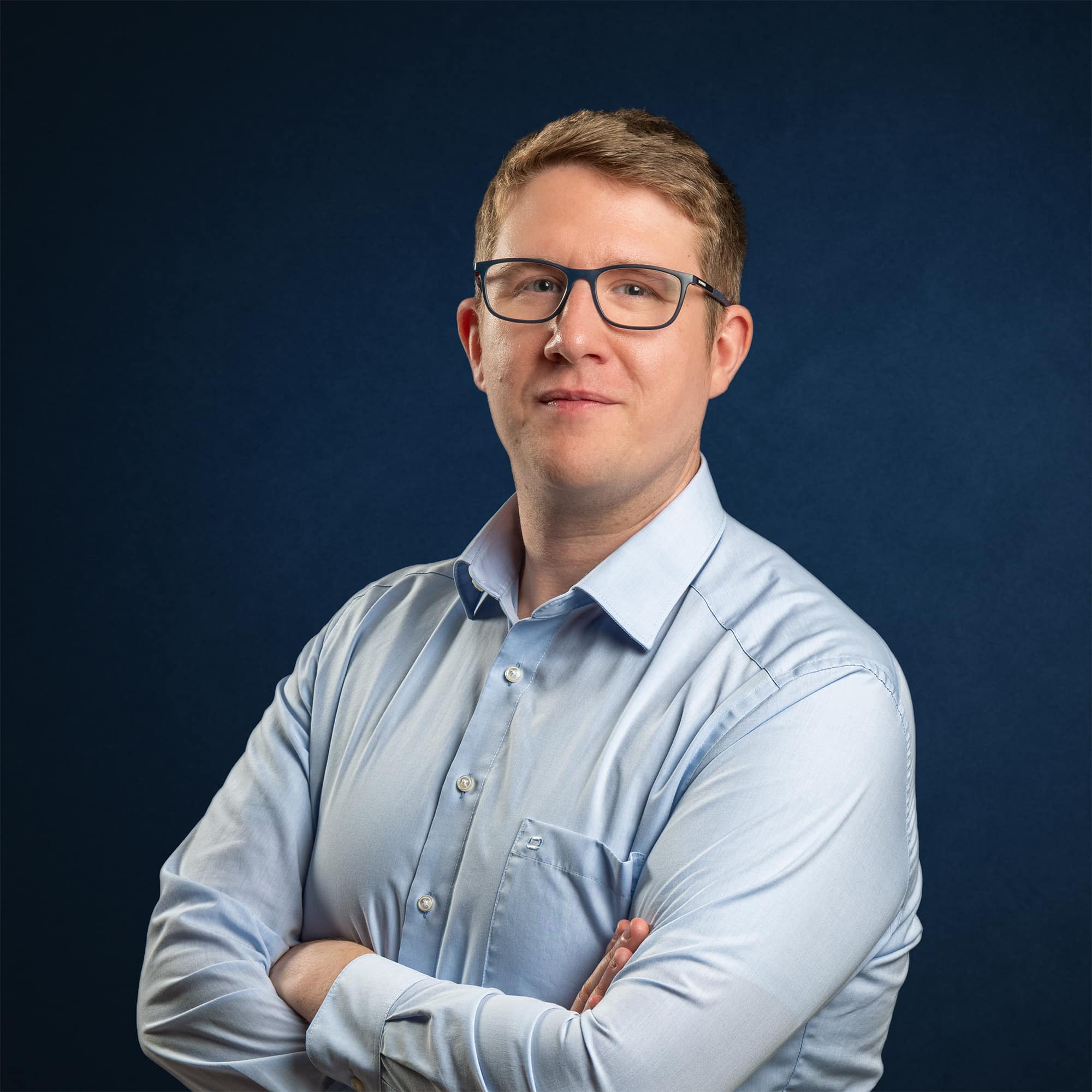 Andreas Klotz, LL.M. blickt mit Brille und verschränkten Armen in einem hellblauen Hemd vor dunkelblauem Hintergrund ruhig in die Kamera.
