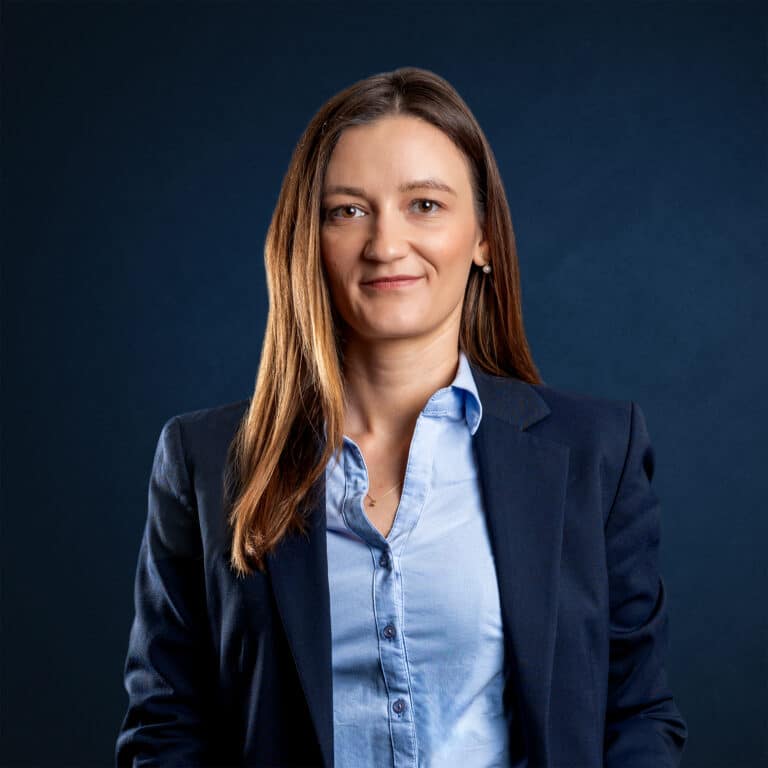 Isabella Köhler blickt in einem dunkelblauen Blazer und hellblauen Hemd vor dunkelblauem Hintergrund ruhig in die Kamera.