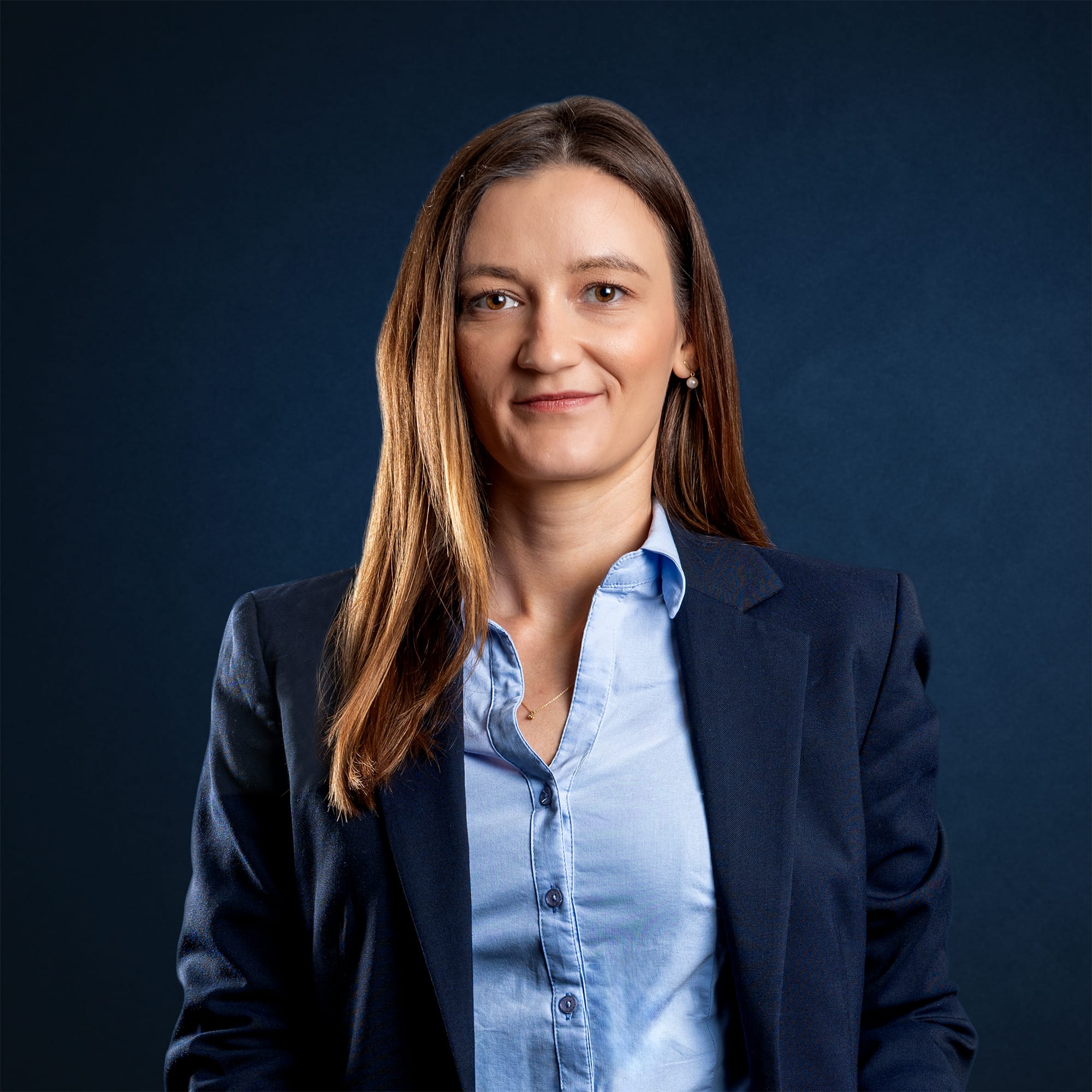 Isabella Köhler blickt in einem dunkelblauen Blazer und hellblauen Hemd vor dunkelblauem Hintergrund ruhig in die Kamera.