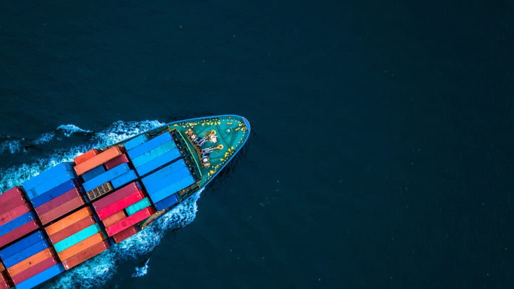 Ein Containerschiff fährt aus der Vogelperspektive durch dunkles Wasser, beladen mit bunten Frachthäfen, und zieht eine helle Bugwelle hinter sich her.