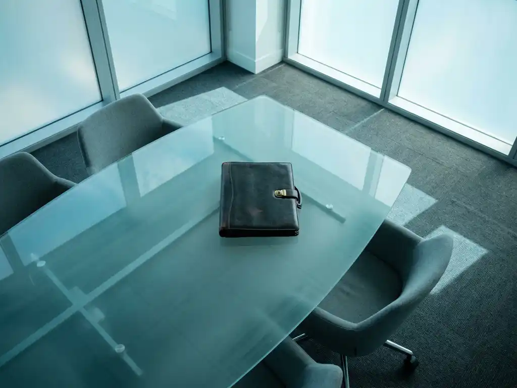 Geschlossenes Lederdossier auf einem gläsernen Konferenztisch mit cyan-blauem Lichtreflex in einem minimalistischen Boardroom.