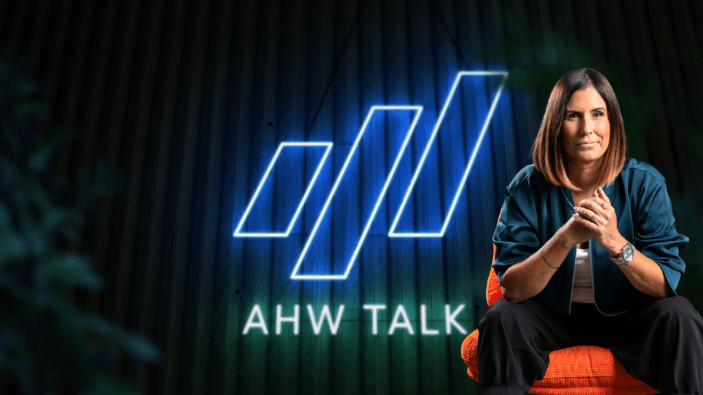 Eine Frau sitzt vor dem leuchtenden Schriftzug „AHW TALK“ und dem blau illuminierten AHW-Logo auf dunklem Hintergrund.